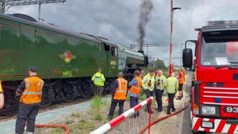 A green locomotive stopped on railway tracks. There is a fire engine to the side. There is black smoke coming from locomotive. Several men in high vis yellow and orange jackets and stood in between the fire engine and locomotive.