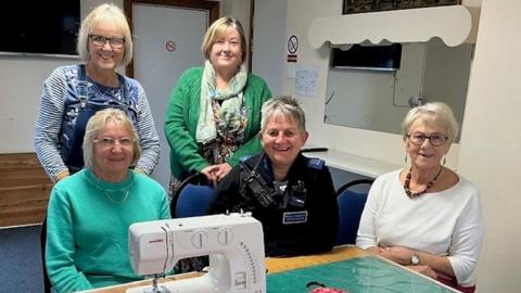 Three women are sat down, with two more women stood behind them. There is a sewing machine in the foreground. There is a police officer sat in the middle. Each person is smiling. 