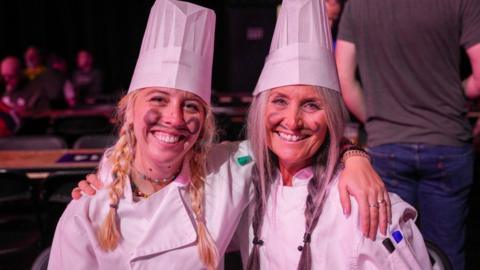Two women wearing a white hat and white top are smiling at the camera. They each have an arm on the shoulder of the other woman. Someone in a grey top has their back to the camera on the right.