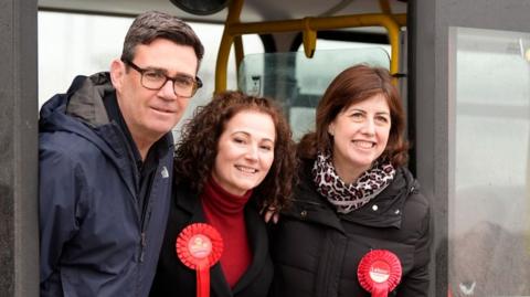 Andy Burnham at a Labour campaign event with its candidate Angeliki Stogia and Deputy Leader Lucy Powell - all three are standing in the entrance of a campaign bus, with the two women wearing red rosettes