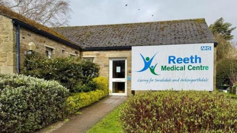 A GP practice which is a single storey building with a glass door and hedges around the entrance. A sign reads Reeth Medical Centre. 