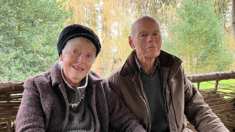 Coral and Ernest Hoyos sit in a small wooden hut behind them are mature trees in a range of greens, oranges and browns. They are in their 80s and Coral is wearing a black hat and purple coat. Ernest is wearing a brown coat.