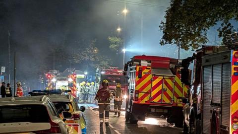 Four fire engines and a police car are parked in a line near the fire in Christchurch. It is dark and the vehicles have their flood lights and flashing beacons switched on. About six or seven firefighters and a couple of police officers can be seen - some are carrying out tasks while others are talking.