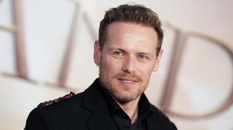 Outlander star Sam Heughan with a stubbly beard and short brown hair in a dark jacket
