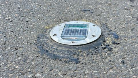 A close up of a grey concrete road. In the middle is a silver metal disk with a clear plastic square.