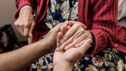 A close-up of a nurse's hands holding those of an an elderly woman wearing a red cardigan and floral dress