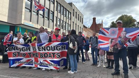 A crowd of people holding Union flags on a pedestrianised street. The largest flag is printed with the words "Enough is Enough, Stop the boats, Patriots of Gloucestershire". One man holds a Union flag with King Charles II printed on it.  