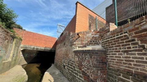 A river with a high brick wall to each side and a red brick bridge over the top. 