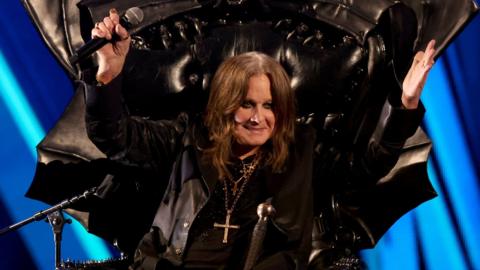 Ozzy Osbourne sat in chair with microphone and sunglasses on.