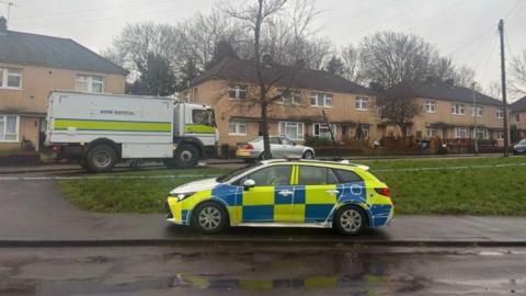 A police car is parked outside a row of beige houses next to a green. The sky is grey and the street is wet.