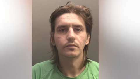 Ryan Burgess looks at the camera, he has shoulder length fair hair and a faint moustache 