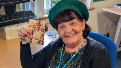 Erika Slater smiles and holds a photo of herself when she was younger and wearing her nurse's uniform. She is sitting at a desk and has a dark blue cardigan and patterned green and yellow top, wearing a green beret.