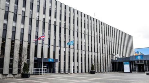 The outside of Darlington Town Hall which is a large, brutalist building with columns of narrow windows. There is a Union Jack flag flying outside the entrance along with another blue flag. 