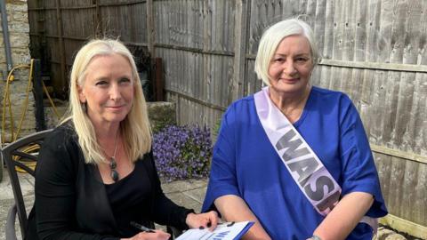 Two women are sat down on chairs in a garden. They are both smiling at the camera. One women has blonde hair and it wearing a black top and jacket. She is holding a clipboard with a piece of paper that has a large heading that reads 'Waspi'. The other women has short grey hair and is wearing a blue top. She is wearing a pink sash that says 'Waspi' on it. 