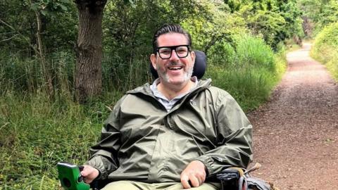 A man with dark hair and glasses wearing a green coat is sat in a wheelchair using a path that is lined by long grass and trees.