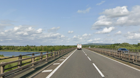 A bridge on a stretch of two-lane motorway passes above a river in the Gloucestershire countryside on a sunny day. There is a lorry travelling in the outside lane with cars coming in the opposite direction on the other carriageway.