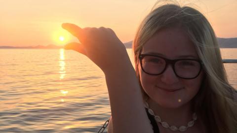 Lucy is wearing glasses and a necklace, posing as if she is trying to pinch the sun at sunset