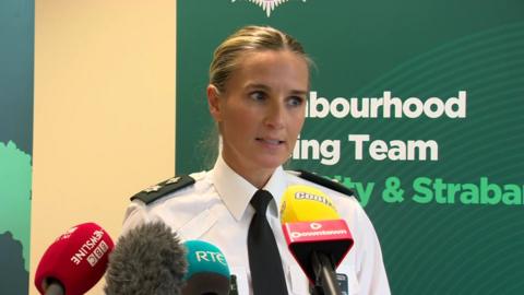 Gillian Kearney speaking at a police press conference. There is a green PSNI banner behind her and four microphones in front. She is in uniform and has blonde hair, which is tied back.