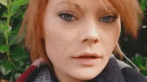A selfie of Kimberly Milne. who has blue almond-shaped eyes, freckles and strawberry blonde hair, which is tied up. She is wearing a scarf and standing in front of greenery.