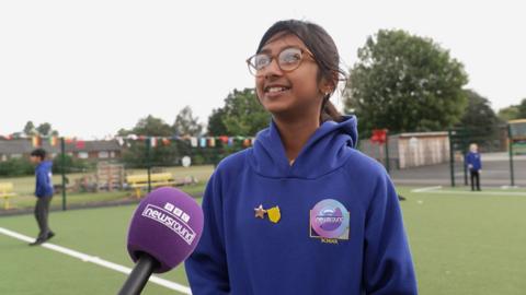A young girl wearing her blue school uniform with badges on her jumper speaking on a football pitch