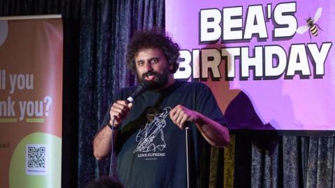 Nish Kumar on stage during the comedy event. A banner reading 'Bea's Birthday' is displayed behind him.
