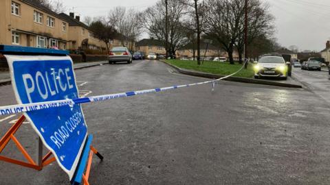 A police cordon is blocking off a residential road with beige houses lining the street. A police car is parked next to the road.