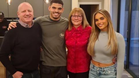 Brian Gibbs (l) in a dark jumper and shirt in a kitchen with his grandson in a brown top and his wife, grandmother Julie, in a red shirt and Gibbs-White's girlfriend Britney in a beige cropped top. Gibbs-White has his arm around his family.