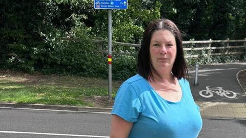 A woman wearing a sky blue T-shirt and blue jeans, standing next to a road with a cycle path on the opposite side of the road