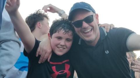Rev Huw Thomas has a black baseball cap, shades and a black Fred Perry shirt and his son has a black T-shirt with a red logo on it. The pair have their arms over each other and are smiling.