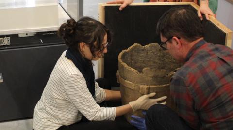 A woman is crouching besides the vessel. She is weather latex gloves and is assisted by a colleague who is also holding the vessel and a third person is holding a container for the artefact.