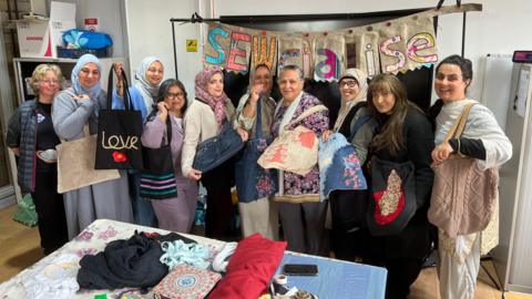 A group of ladies stand in front of a big sign which says 'Sewcialise'. The sign is made of handmade textile bunting, and the women are all holding up tote bags made of different materials. In front of them is a table full of rolls and piles of colourful textiles, boxes of thread and other related items.
