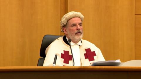 Judge Lord Beckett in court, wearing a white robe with red crosses and a judge's wig. 