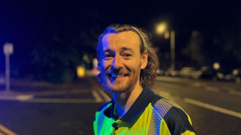 Eddie, a Glasgow street cleaner, who cleans the city's streets during the night. He is smiling at the camera and wearing a yellow. hi-viz jacket.