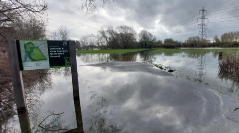 A view of flooded Oatlands & King George's Recreation Ground in Oxford. The sign with the name of the park is in vision. The rounds are saturated. It is overcast.