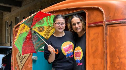 Two women look out of a brown caravan. The woman of the left is holding a colourful umbrella