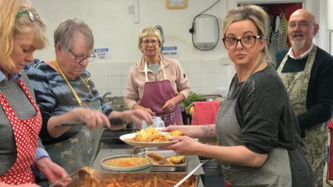 Four women and a man dressed in aprons serving up a pasta bake with garlic bread. A woman is seen at the front of the picture holding a plate.