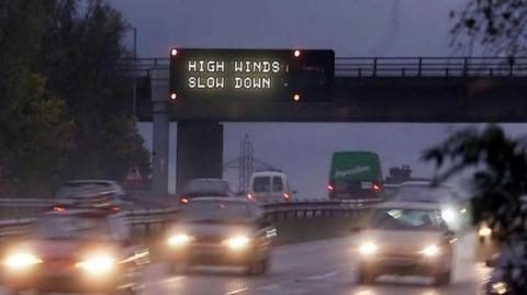 An illuminated matrix sign above a busy motorway at night warns of high winds and to slow down