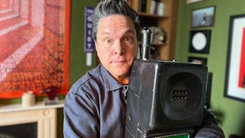 Dom Joly is looking at the camera holding the giant mobile telephone famous for one of his sketches on Trigger Happy TV. He is inside a house with art framed and hung on the walls.