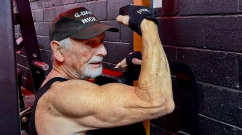 Mick Wadge flexes his right arm while using a weight-training machine at the gym.