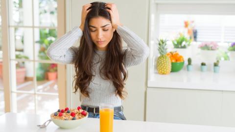 A woman sits at a breakfast bar with a bowl of cereal and a glass of orange juice. She is holding onto her head as if in pain. 