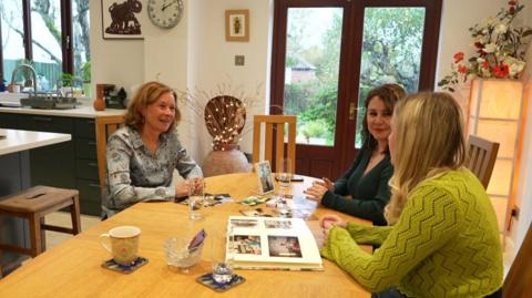 Three women sit around a kitchen table talking, in the middle of the table are a collection of photographers of their late mothers