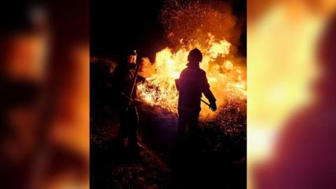 Two firefighters seen battling the gorse fire at night. The area around them is very dark and only their silhouette is visible. In front of them is a large fire.