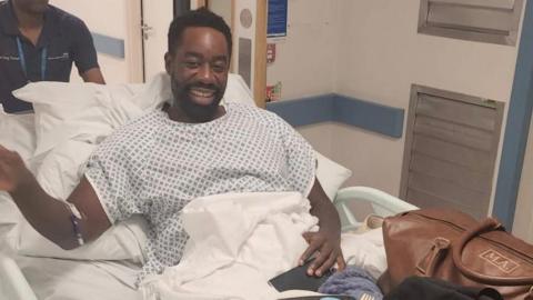 Matthew Allick in a hospital robe and on a stretcher inside a hospital. He smiles at the camera.