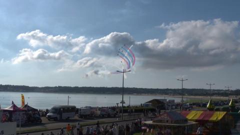 The Red Arrows flying over the sea in a V formation with red, white and blue smoke trailing from them while two jets fly around them. There are trade stands and visitors in the foreground on the St Helier waterfront