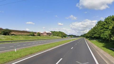 An empty A417 Brockworth bypass with grass and trees on the verges on a sunny day.
