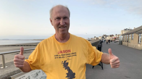 Jason Vickers in a yellow marathon t shirt as he trains in Burnham. He has both thumbs up as he smiles at the camera. In the background, you can see the beach road in Burnham.