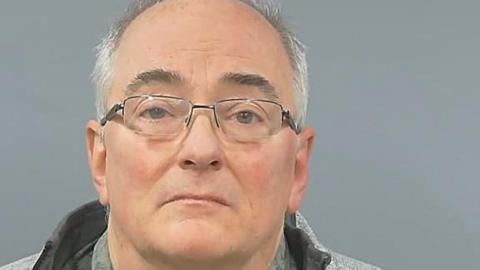 Mugshot of an older man in glasses and a grey jacket