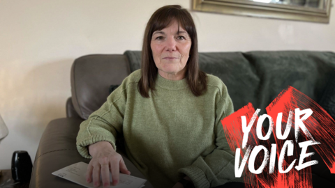 A woman with shoulder-length dark hair, wearing a green jumper, sits on a sofa looking at the camera