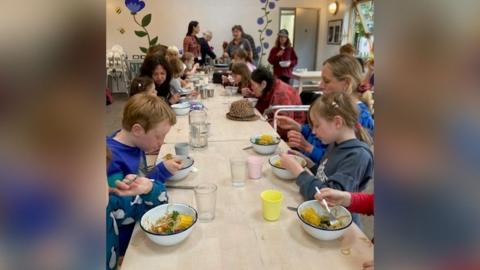 Children sitting around a long wooden table, eating vegetables from white bowls.