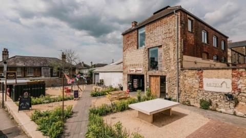 A renovated brick building with redeveloped garden out front with tables and greenery and an old great corrugated building next to it